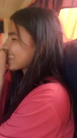 A young woman with long dark hair wearing a bright pink t-shirt is visible in the video. She is in the passenger seat of a moving vehicle, and turns her head to look directly at the camera. The driver, a man wearing a red baseball cap, is visible in the background. The environment appears to be inside a car with bright sunlight. The woman appears to be talking or singing, and smiles at the camera.