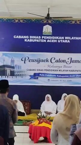 The video captures an indoor gathering, likely a "Peusijuek" ceremony, which is an Acehnese blessing ritual, for prospective Hajj pilgrims. The event is hosted by the Regional Education Office of North Aceh Regency.

The scene is set in an auditorium, indicated by the main blue banner at the back that reads "CABANG DINAS PENDIDIKAN WILAYAH KABUPATEN ACEH UTARA" (Regional Education Office, North Aceh Regency). A smaller, more decorative banner below it announces the specific event: "Peusijuek Calon Jamaah Haji Keluarga Besar CABANG DINAS PENDIDIKAN WILAYAH KABUPATEN ACEH UTARA" (Peusijuek for Prospective Hajj Pilgrims, Extended Family of Regional Education Office, North Aceh Regency). This banner features an image of the Kaaba, a significant symbol in Islam, and specifies the location and date as "Aula Cabdisdik Kabupaten Aceh Utara" (Auditorium of Regional Education Office, North Aceh Regency), "Lhokseukon, 30 April 2028".

At the front of the hall, on a raised platform, a group of women are seated, predominantly dressed in white modest attire and headscarves. In front of them, on a low table, are items covered with red and yellow cloths, characteristic elements of traditional Acehnese ceremonies.

The video is filmed from the perspective of an attendee in the audience. Several individuals, both men wearing songkok (Islamic caps) and women in hijabs, are visible in the foreground, facing the front and attentively observing the proceedings. The atmosphere appears solemn and respectful. Islamic chanting or prayer is audible, contributing to the spiritual nature of the event. A ceiling fan is visible above, suggesting an indoor venue. The event is taking place in Meunyematangubi, Indonesia, within the North Aceh Regency as indicated by the banners.