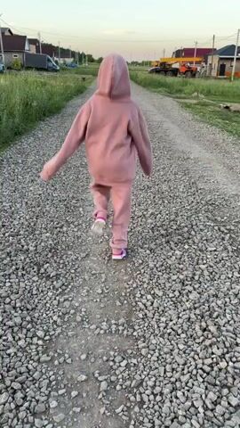 A young child wearing a pink hooded sweatshirt and pants walks away from the camera down a gravel path. The path is flanked by grass and houses on either side. In the background, there are some construction vehicles. The sky is a pale blue with hints of pink and orange, suggesting it is either sunrise or sunset.