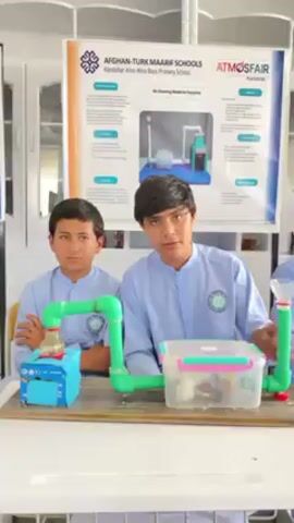 Three young boys, likely students from Afghan-Turk Maarif Schools in Kandahar, are presenting a science project indoors. They are dressed in light blue, long-sleeved uniforms with a school emblem on the chest.

The project, displayed on a white table, is an "Air Cleaning Model for Factories." It features a blue box with a red-capped bottle, connected by green PVC pipes to a clear plastic container filled with various filtering materials. Another green pipe extends upwards from the container, topped with a funnel-like structure.

Behind the boys, a poster board reinforces the project's details, stating "AFGHAN-TURK MAARIF SCHOOLS," "Kandahar Aino-Mina Boys Primary School," and "ATMOSFAIR," alongside an illustration and descriptive text. One boy is actively speaking and gesturing towards the model, while the other two observe attentively. The setting appears to be a brightly lit classroom or science fair hall, with white furniture visible in the background.