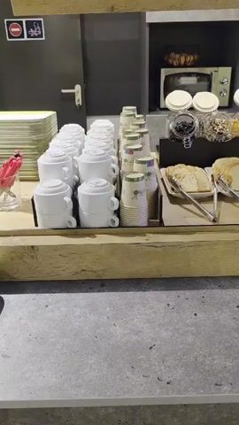 The video shows a breakfast buffet station in what appears to be a hotel or similar establishment. The station is made of light-colored wood and features several shelves. On the top shelf is a black bowl with apples inside, as well as a microwave. The main counter of the station features rows of stacked white mugs, paper cups, jars filled with toppings like dried fruit and nuts, and platters of sliced white bread with tongs. To the right, there are containers of cheese, ham, and small yogurt cups. The floor is a gray tile.  There are no people visible in the video. The camera pans slowly to the right.