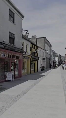 The video shows a pedestrianized street lined with buildings. On the left, there are storefronts with awnings. A white A-frame sign is visible in front of one of the shops. The street is paved with light-colored stones, with a decorative border of darker stones running along the edges. Several people are walking down the street in the distance. The sky is cloudy.