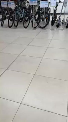 The video shows the interior of a retail store, possibly a department store or sporting goods store, featuring a selection of bicycles and electric scooters displayed on a tile floor. The bikes are arranged in rows, some standing independently and others racked on metal stands. Each appears to have a price tag or information card attached. Several people are walking in the background, appearing to be customers browsing.  The store's lighting is bright, and there are windows and displays of clothing and jewelry visible in the background.