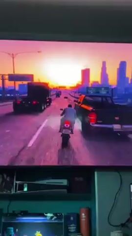 A video game is being played on a television. The game features a character riding a motorcycle on a highway as the sun sets over a city. The highway is populated with other vehicles. The game is vibrant with a purple and orange color scheme. Below the television is a shelf with a PlayStation 5 console, a clock, and a few other items.