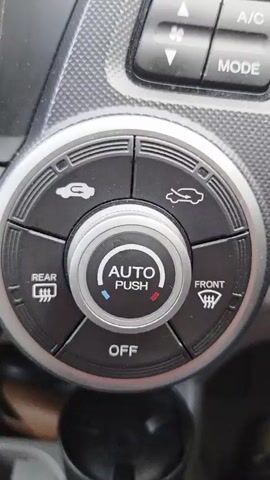 The video shows a close-up of a car's climate control panel. The panel is circular with a central dial that has "AUTO" and "PUSH" printed on it, surrounded by a red and blue indicator light. Around the central dial are five buttons: one with a car and an upward arrow, another with a car and a downward arrow, one labeled "REAR" with a defroster symbol, one labeled "FRONT" with a defroster symbol, and one labeled "OFF". The buttons are black with silver icons and text. The surrounding panel is dark gray and textured. The lighting suggests the car is in a well-lit environment, possibly daylight. There are no people or other objects visible in the frame. The video pans slightly and zooms in on the climate control panel, focusing on the details of the buttons and dial.