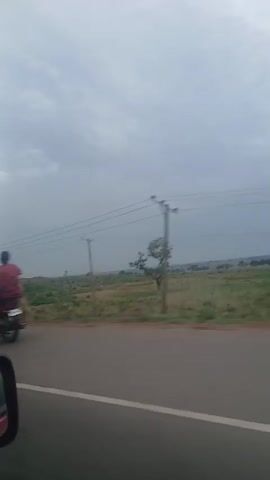 The video shows a view from a car driving on a paved road. A red moped passes by in the beginning, the driver wearing a hat. The roadside is a mix of green shrubbery and reddish-brown earth, with scattered small trees. Power lines stretch across the sky, and the sky is overcast with gray clouds. The car's side mirror is visible in the lower left corner of the frame.