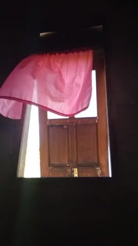 A pink curtain is attached to the top of a window. The curtain is blowing in the wind. Outside the window is a wooden door.