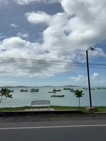 The video shows a bay with several fishing boats at anchor. The water is calm and the sky is partly cloudy. In the foreground, there is a road with a sidewalk and a patch of grass. A white bench is visible on the grass, as well as a wooden signpost. A utility pole with wires is also present. The camera pans from left to right, revealing more of the bay and the boats. A tree is visible on the left side of the frame, and on the right side, there is a small plant. A silver van is parked on the right side of the road, partially obscuring the view of the bay. The overall atmosphere is peaceful and serene.