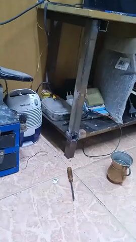 Here's a concise, forensic description of the video:

The video shows a cluttered indoor space, likely a workshop or storage area. The floor is covered with patterned ceramic tiles. The main subject is a metal desk or workbench, constructed with welded square tubing. The desk is partially covered with various objects, including electronic components, wires, and containers. 

To the left of the desk is a small blue toolbox and a white air purifier unit labeled "Pro Breeze". In front of the desk on the floor is a screwdriver and a copper-colored mug filled with liquid. 

The lighting is dim, and the camera is stationary. There are no people visible in the shot. The focus is on the desk and the surrounding objects, suggesting a possible DIY project or a general storage area. There is a buzzing sound throughout the video.