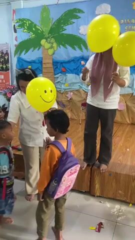 The video shows a group of young children and two adult women in what appears to be a classroom or event space decorated with a beach theme. On the right, a woman wearing a hijab and a unicorn headband stands on a wooden platform, holding a stick and a yellow balloon with a smiley face. She is interacting with the children. To the left of her, another woman wearing a white top and cat ears is holding a microphone and a yellow balloon. She is also engaging with the children, who are mostly wearing backpacks. One child in a yellow shirt is reaching for the balloon held by the woman with the microphone, while another child in an orange shirt with a purple backpack stands between them. The background features a mural of a beach scene with palm trees and water. The overall atmosphere suggests a children's activity or event.