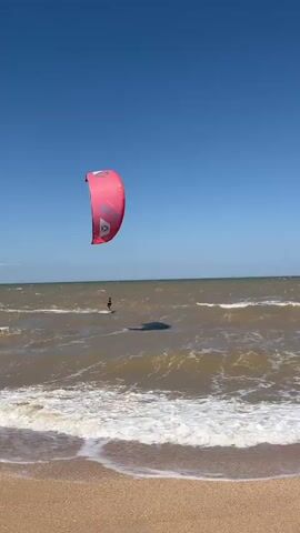 A person kitesurfs on choppy brown water under a clear blue sky. The kitesurfer is on a board, with a large pink kite with a white logo above them. Waves crash on a sandy beach in the foreground. The camera is positioned low on the beach, looking out at the ocean.