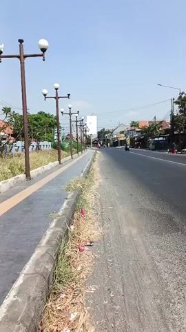 The video shows a sidewalk with lampposts and a street. The sidewalk is paved with gray tiles and has a concrete curb. There is a strip of dry grass and some litter between the sidewalk and the street. The street is asphalt and appears to be a two-lane road. There are buildings and trees on the side of the street. The sky is clear and blue. There are no people visible in the video.