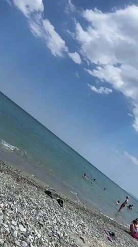 A video shows a tilted view of a beach and the ocean. The beach is covered in pebbles, and the ocean is a calm, turquoise color. Several people are visible swimming and wading in the water. The sky is a clear blue with a few scattered white clouds. The camera appears to be held by someone walking along the shore, resulting in the tilted perspective.
