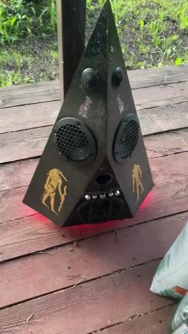 A black, triangular-shaped speaker system with golden Egyptian-themed decals is placed on a wooden deck outdoors. The speaker features multiple speaker grilles, control knobs, and glowing red and yellow lights at its base. The speaker plays music in two separate clips, with the sound changing from music to silence between the clips. In the second clip, the lights at the bottom of the speaker change from red to yellow.
