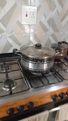 The video shows a stove top on which two pots with lids are sitting. The left pot is larger and filled with boiling water. There appear to be plantain chips inside. The smaller pot next to it is also covered with a lid. The stove top is stainless steel. The walls are light-colored with gray and white geometric tiles.