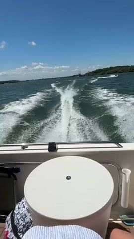 The video shows a first-person perspective from the stern of a moving boat on a sunny day. The boat is creating a white wake that trails behind it across the green-blue water. In the foreground, part of the boat's white stern is visible, including a round, white cooler or lid and a white fishing rod holder on the right. A person's leg and a patterned bag are partially visible on the left. In the mid-distance, another smaller boat or personal watercraft is moving away from the camera on the right, also creating a wake. The background features a distant shoreline with trees, and a clear blue sky with some scattered white clouds. The boat continues to move forward throughout the video.