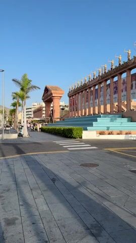 The video shows a wide street with a sidewalk. On the right side of the street is a building with a classical facade, featuring columns and statues. A wide staircase with turquoise steps leads up to the building. Palm trees are visible along the street, and there are some shops and buildings in the background. Several cars and a taxi are driving on the street. A few people are walking on the sidewalk and crossing the street at a crosswalk. The sky is clear and blue, suggesting a sunny day. The overall scene appears to be a commercial or tourist area.