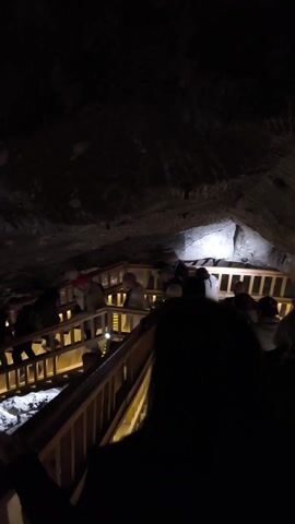The camera is oriented vertically, looking down into a narrow, dimly lit passageway between rock walls.  A wooden staircase with multiple landings descends through the space. People are walking down the stairs. The space is narrow and constrained.  The walls appear natural and uneven, suggesting a cave or grotto. Some lighting fixtures are attached to the staircase structure.  The camera is unsteady, contributing to a sense of disorientation. The foreground is obscured by a dark object, possibly the person holding the camera.
