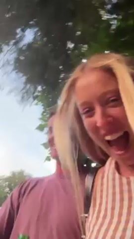 Two people, a blonde woman and a man with dark hair and a pink shirt, are walking outdoors. The woman is filming, and the camera is shaky. The man is holding a green bottle. They are both laughing. Trees and sky are visible in the background.
