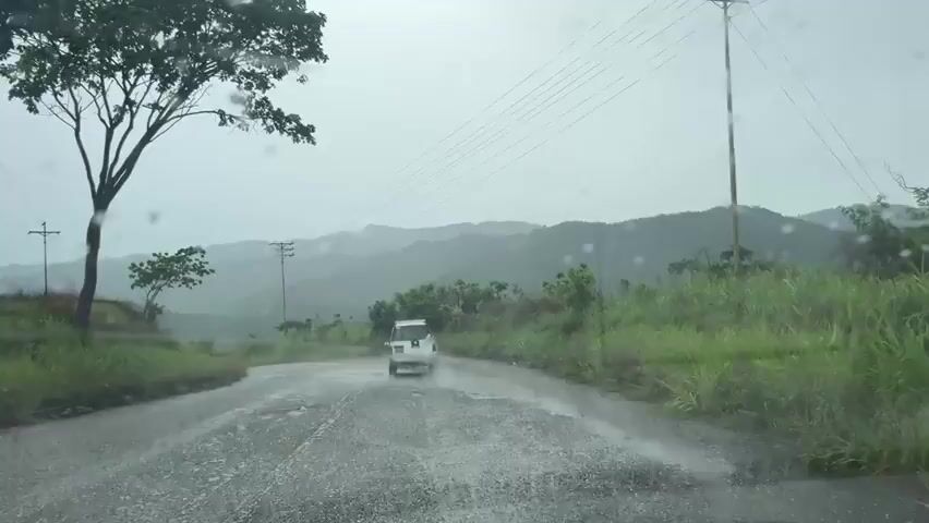 The video, filmed from inside a moving vehicle through a rain-streaked windshield, shows a white SUV driving away from the camera on a wet, paved road with potholes. The road curves slightly to the left. On both sides of the road, there is lush green vegetation, including tall grass and trees. Utility poles and power lines run parallel to the road on the left side. In the background, rolling green hills and mountains are visible under a cloudy, overcast sky, indicating rainy weather. The raindrops on the windshield become more prominent as the video progresses.