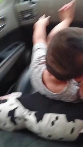 The video shows a young child seated on a black leather car seat, viewed from behind. The child is wearing a light gray t-shirt. A person's arm, with light skin tone and visible fingers, rests on the child's lap. The person's hand is also visible near the bottom of the frame. Beneath the child and person, a pillow with a black and white dalmatian-like pattern is visible. The background suggests the interior of a car, with a dashboard and a screen visible on the right side. The lighting is low, indicating it could be evening or a dimly lit environment. There is no visible text. The overall scene appears to be a candid moment of a child in a vehicle, possibly on a journey.