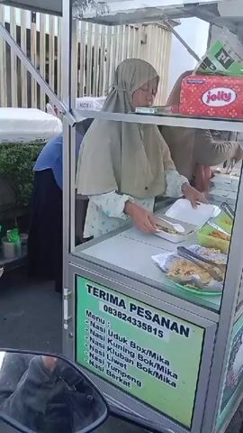 The video shows a woman wearing a hijab preparing food in a street food cart. The cart has a glass display case and a sign that reads "TERIMA PESANAN" (We accept orders) in green and white lettering, followed by a phone number and a menu list of various rice dishes. Inside the cart, there are white styrofoam containers and various food items. Another person is visible in the background, also working in the cart. The cart appears to be parked outdoors, with a fence and foliage visible in the background. The overall scene depicts a food vendor preparing orders.
