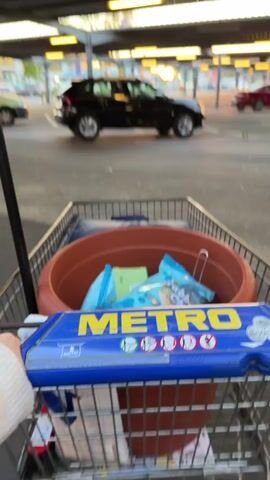 The video shows a person pushing a Metro shopping cart across a covered outdoor parking lot. The cart contains a large brown terracotta pot, two blue bags of chips (one with "Bubble Gum" partially visible, the other with "Chicken"), a small green rectangular item, and some blue fabric or jeans. In the background, several cars are parked, including a black SUV moving slowly and a red car. The lighting suggests late afternoon or early evening, with warm sunlight coming from the side. The "METRO" logo is clearly visible on the shopping cart handle. The setting is likely a Metro Cash & Carry store in Sankt Augustin, Germany.