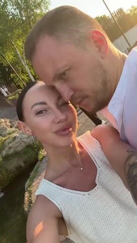 A couple is taking a selfie together outdoors. The woman is wearing a white knitted tank top, a silver necklace, and hoop earrings. The man is wearing a light pink collared shirt and has a tattoo on his arm. They are standing on a stone path near a small body of water and some greenery. There are other people in the background, including a woman in a striped shirt. The man kisses the woman on the forehead. The sun is setting, casting a warm glow on the scene.