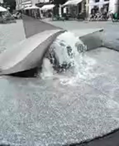 A close-up shot of a modern fountain in a public space. Water erupts from a central point, creating a turbulent cascade. The fountain's design appears abstract, with large, smooth, angular stone elements surrounding the water source. In the background, blurred buildings and awnings suggest an urban setting, with hints of people and vehicles in the distance. The ground surrounding the fountain is paved with a speckled stone material. The video is a static shot, with the only movement being the water flow.