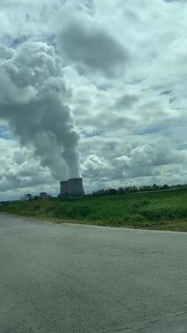 The video shows two cooling towers of a power plant venting steam into the atmosphere. The structures are gray, and the steam plumes merge into a cloudy sky. The foreground features a paved road and a grassy field, providing a sense of distance to the power plant. The camera appears to be moving slowly past the location, perhaps from a car. The overall scene is calm and neutral, capturing an industrial landscape.