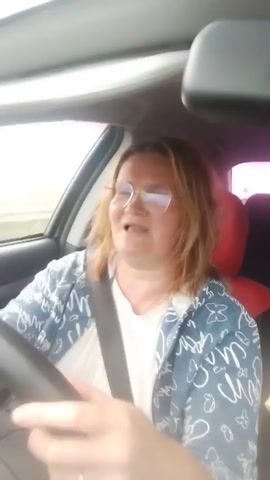 A woman with blonde hair and glasses is driving a car. She is wearing a blue and white patterned jacket over a white shirt. Her mouth is open as if she is speaking or singing. The car appears to be moving, with a blurred view of the outside environment visible through the window.