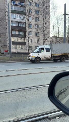 The video shows a vehicle moving along a road, passing a white truck with a covered bed and a flatbed trailer. A small orange utility vehicle with two workers operating it is visible on the side of the road. One worker in a dark coat and another in protective gear are next to the utility vehicle. The setting appears to be an urban environment with buildings and trees lining the road. Tram tracks are visible on the road surface.
