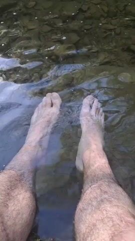The video begins with a first-person view of a person's bare feet submerged in a shallow, clear stream. The water gently flows around the feet, and the stream bed is visible beneath the surface, with various rocks and pebbles. The person is standing. The camera then pans up to reveal a waterfall cascading into the stream. The waterfall is surrounded by lush greenery and trees. The sun shines brightly, creating a glare on the water. The scene is natural and peaceful.