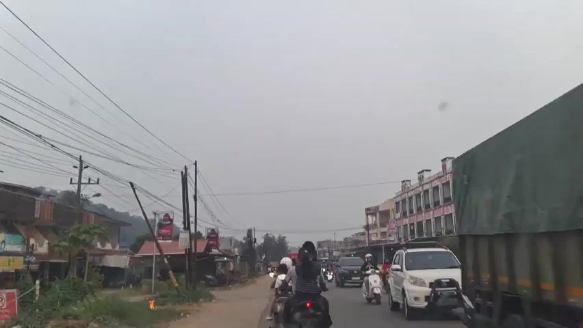 The video shows a street scene with several motorcycles and cars. The weather appears to be overcast. On the left side of the road, there are several small buildings and utility poles with many wires. On the right side of the road, there is a multi-story building with a row of windows. Several people are riding on the motorcycles, and at least one person is in a car. The vehicles are moving down the road. There is a white SUV in the foreground on the right.