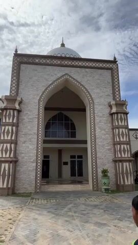 A group of students are gathered outside a mosque. The mosque is a large, light-colored building with a dome and intricate archways. The students are mostly wearing green shirts, and some are carrying bags. Two boys in the foreground are walking away from the camera, towards the mosque. The sky is cloudy.