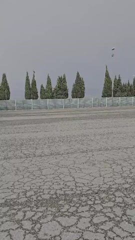 The video pans across a cracked asphalt lot, possibly a parking area or industrial site. In the background, a line of tall, slender evergreen trees borders a white fence. Above the trees, several small, dark objects are visible in the hazy sky, appearing to be either birds or possibly drones. Further into the lot, a row of parked semi-trucks is visible, varying in color and size. The sky is overcast, suggesting a cloudy or possibly foggy day. The overall mood is subdued and somewhat desolate, with no people visible. The location is identified as Épila, Spain.