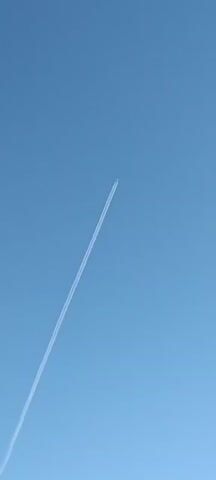 This media file captures an airplane leaving a prominent contrail against a clear blue sky. The airplane itself is a small, white object at the apex of the contrail, appearing as a speck against the vast expanse of the sky. The contrail is a long, white, and well-defined trail that stretches diagonally across the frame from the lower left to the upper right.

The scene is an outdoor setting, specifically the sky. The weather appears to be clear with no visible clouds, indicated by the uniform blue color of the sky. The bright, uniform lighting suggests it is daytime, likely midday, given the absence of strong shadows and the intensity of the blue. There are no identifiable location cues beyond the general sky. The overall emotion conveyed is one of vastness and perhaps a sense of travel or distant movement.

There is no visible text in the media file. The focus is solely on the airplane and its contrail against the sky.
