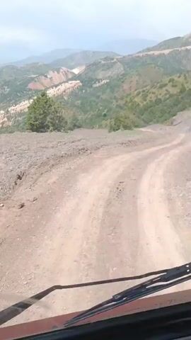 The video shows a scenic view of a mountainous landscape in Nurekata, Uzbekistan. A dirt road winds through the terrain, and the camera appears to be mounted inside a vehicle, possibly an SUV, as indicated by the windshield wipers visible at the bottom of the frame. The mountains are covered in a mix of green vegetation and exposed, reddish-brown soil, suggesting a semi-arid or mountainous climate.

In the background, a cable car can be seen suspended between two poles, traversing a steep slope. The cable car itself is dark and has a green light illuminated. The sky is overcast with some patches of lighter clouds, suggesting it might be midday or late afternoon. The overall impression is one of natural beauty and ruggedness, with the cable car hinting at human activity and infrastructure in this otherwise wild setting. The activity taking place is a journey through this mountainous area, potentially for tourism or access to remote locations. There is no visible text in the media file.