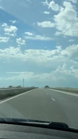 The video shows a view from inside a car driving down a straight highway. The road is grey asphalt with white lane markings and guardrails on either side. Beyond the guardrails, there's green vegetation. In the distance, an electrical tower stands on the left side of the road. The sky is blue with fluffy white clouds, and there are hints of sunlight. The car's dashboard is visible at the bottom of the frame.