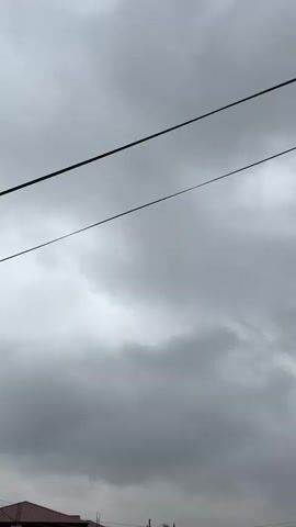 The video shows a cloudy sky above a partially built brick wall. There are power lines running across the top of the frame. Some tall grasses and palm trees are visible behind the wall.  The overall impression is of a somewhat gloomy, possibly overcast day.