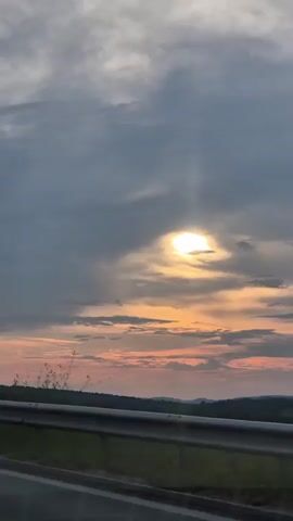The video captures a scenic view of a sunset over a rural landscape in Peshtera, Bulgaria. The primary subject is the sky, dominated by dramatic clouds that partially obscure a bright, radiant sun. The sun is seen peeking through a break in the clouds, casting a warm, golden glow across the sky. Streaks of orange and pink are visible along the horizon, indicative of the sunset.

In the foreground, a highway guardrail runs horizontally across the frame, suggesting the view is from a moving vehicle or a roadside vantage point. Beyond the guardrail, a strip of green land stretches out, leading to a low-lying horizon marked by distant hills or mountains. The overall atmosphere is serene and peaceful, with the vibrant colors of the sunset evoking a sense of natural beauty and tranquility.

The time of day is clearly sunset, as evidenced by the low position of the sun and the characteristic colors in the sky. The weather appears to be partly cloudy, with the clouds playing a significant role in the visual drama of the scene. The location context of Peshtera, Bulgaria, adds a specific geographical element to the landscape. No people or specific objects, other than the guardrail, are clearly identifiable in the video. There is no visible text.