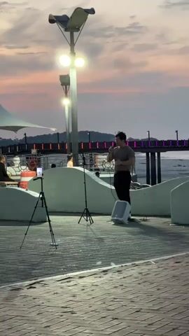 This media file captures a street performer singing into a microphone while standing on a paved walkway. The performer is a young man, dressed in a grey t-shirt and black pants, with a portable speaker next to him. Two tripods with cell phones are set up in front of him, suggesting he is also recording his performance.

The scene is set outdoors during sunset, as indicated by the warm, colorful sky painted with hues of orange, pink, and purple. The setting appears to be a waterfront promenade or pier, with a body of water visible in the background. A pier with colorful lights is also present in the distance. Streetlights, illuminating the walkway, are visible, casting a bright light on the performer. Other people can be seen in the background, suggesting a public gathering or a lively area.

The atmosphere appears to be calm and pleasant, with the performer engrossed in his singing. The overall mood is one of relaxed entertainment, with the beautiful sunset adding to the ambiance. There is no visible text in the media file.