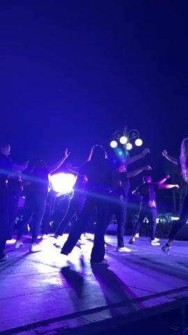 A group of about 10 dancers perform on an outdoor stage under blue stage lighting. The dancers, who appear to be both male and female, are all wearing dark tops and pants. Some are wearing white shoes. Their faces are not clearly visible, as the video is shot from the back, but they are actively moving in a choreographed routine. The stage floor is illuminated with purple and white light. In the background, a crowd of people can be seen, and above the stage, there's a lamppost with multiple bulbs. The sky is a clear dark blue. The overall impression is of a nighttime public dance performance.