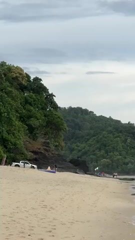 The video shows a wide shot of a beach with a calm, shallow ocean. The sand is light brown and stretches across the foreground. In the distance, a lush, green forested hill rises behind the beach. The sky is overcast with grey clouds.

On the beach, there are several people walking and standing. Some are near the water's edge, and others are further back. There are a few vehicles parked on the sand, including a white SUV. There are also some watercraft visible, including what appear to be kayaks or canoes and a yellow boat.

The ocean is very shallow, with the water only coming up to people's ankles in most areas. Small waves are gently lapping the shore. There are some exposed rocks and sandbars in the water. The overall impression is a peaceful, somewhat crowded beach scene on a cloudy day.