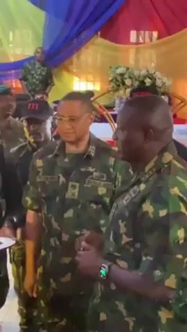 The video shows a group of military officers in camouflage uniforms standing together. They appear to be in an indoor event space, decorated with yellow, blue, and red drapes. One officer is handing something small to another officer who is wearing glasses. Other officers are standing around them, some with hats and insignias. They are conversing and listening attentively. There is a table with flowers in the background.