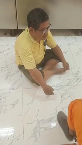 The video shows a man sitting cross-legged on a tiled floor. He is wearing a yellow polo shirt and olive-green shorts. He has short, dark hair and is wearing glasses. He appears to be talking to someone off-camera. Part of another person's legs, wearing orange pants and a grey shoe, is visible in the lower right corner of the frame. The background consists of white tiled walls with some metallic cabinets or appliances. The lighting appears to be artificial, casting some shadows on the floor. The overall environment suggests an indoor setting, possibly a kitchen or a utility area.