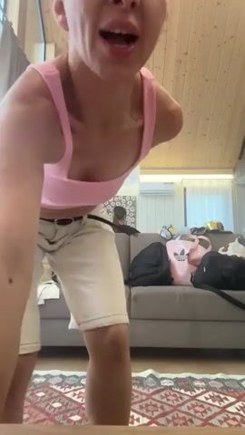 The video features a woman with blonde hair, wearing a pink crop top, white shorts, and white socks. She is in a room with a wooden ceiling and walls. A grey couch with some items on it, including a pink bag with an Adidas logo, is visible in the background. The woman is dancing and appears to be happy and energetic, making hand gestures and forming a heart shape with her hands. The lighting suggests it is daytime.