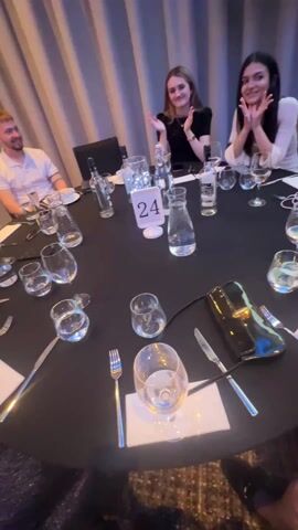 The video begins with a close-up of a formal table setting, complete with a tablecloth, cutlery, glasses, and a table number marked "24". Three people are visible in the background, smiling and clapping. The camera then moves to show a wider view of the room, revealing that it is a crowded banquet hall. There are many people seated at tables, most of whom are looking towards a stage. The room has blue uplighting, and a screen is visible in the distance.