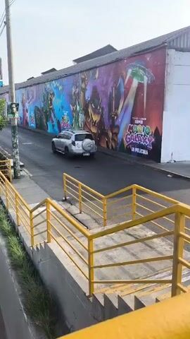 The video shows a roadside scene in a city, possibly in South America. The camera pans from a large mural on a wall depicting characters and imagery from the "Super Mario Galaxy" movie, past a yellow metal railing, to reveal a busy road. Cars and a bus are seen moving along the multi-lane highway. White arrows painted on the road indicate the direction of traffic. On the left side of the road, a large stadium structure is visible, partially covered in protective material. Billboards and power lines are also present in the background. The weather appears to be sunny.