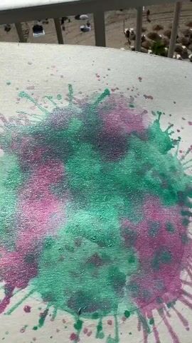 The video displays a close-up of a circular splatter painting with green and pink glitter paint. In the background, a beach scene is visible with people, umbrellas, and the ocean. The camera slowly pans across the painting, revealing the texture and the way the glitter catches the light. The sun reflects off the water, creating a shimmering effect.